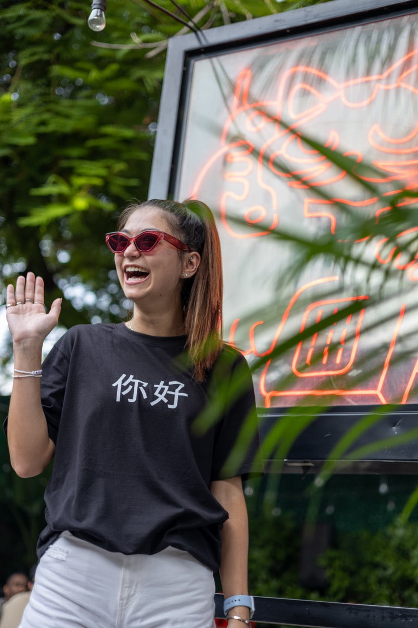 Dani Kott models the 你好 "Nĭhǎo" Hi in Chinese in Pinyin in black Greetings Tee Shirt in front of a neon lucky cat sign from Happy interactions