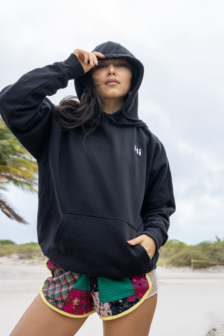 Hi lux embroidered Hoodie in black with Hi waving hand emoji on right sleeve, worn with Hi black socks, modeled by Assiya Mukanova