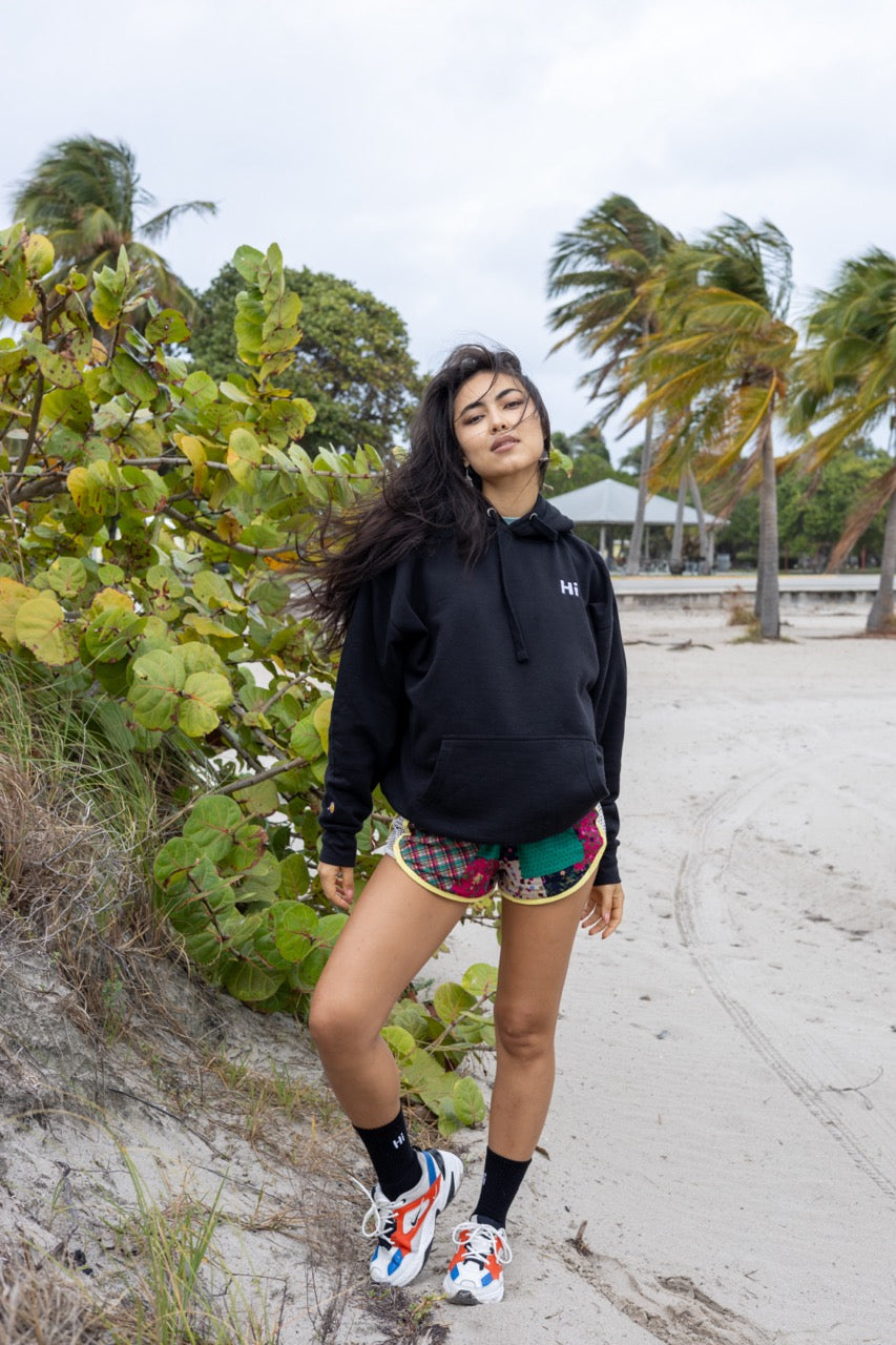 Hi lux embroidered Hoodie in black with Hi waving hand emoji on right sleeve, worn with Hi black socks, modeled by Assiya Mukanova