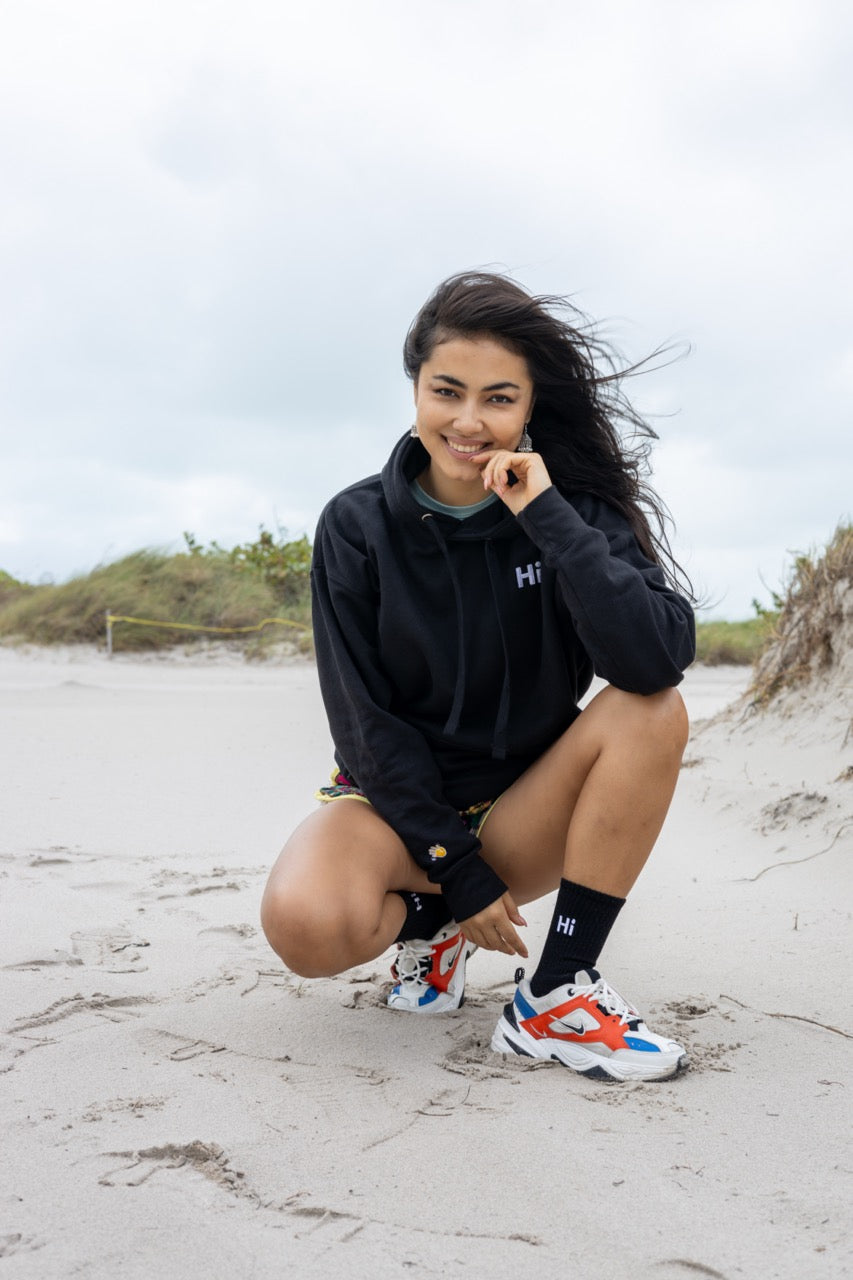 Hi lux embroidered Hoodie in black with Hi waving hand emoji on right sleeve, worn with Hi black socks, modeled by Assiya Mukanova