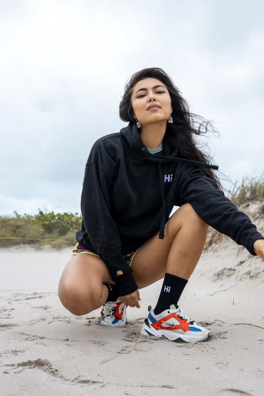 Hi lux embroidered Hoodie in black with Hi waving hand emoji on right sleeve, worn with Hi black socks, modeled by Assiya Mukanova