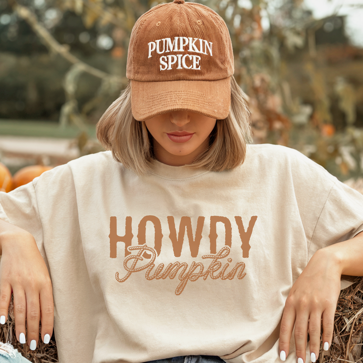 Person wearing a 'Pumpkin Spice' cap and 'Howdy Pumpkin' shirt in an outdoor setting with pumpkins.
