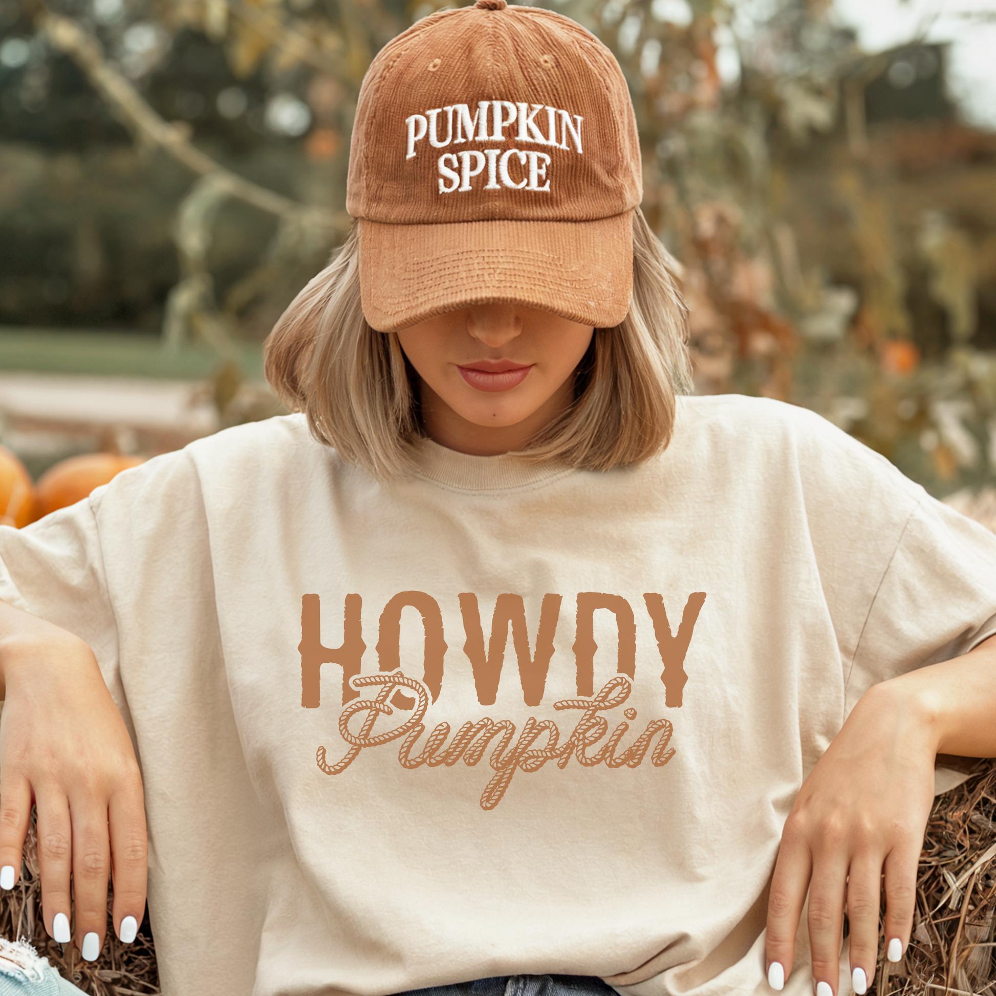 Person wearing a 'Pumpkin Spice' cap and 'Howdy Pumpkin' shirt in an outdoor setting with pumpkins.