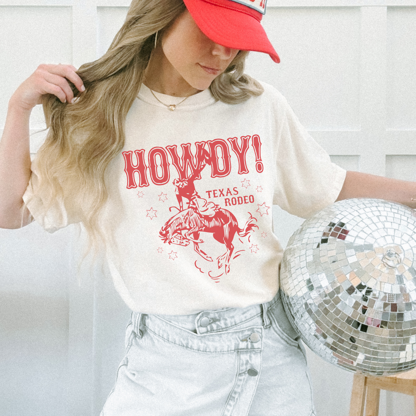Person wearing a 'Howdy! Texas Rodeo' t-shirt and red cap, holding a disco ball.